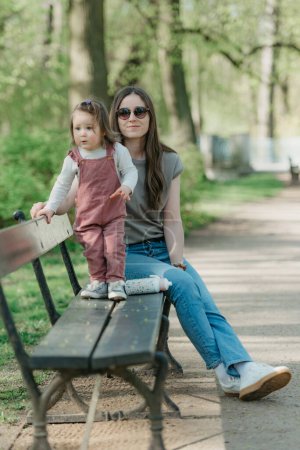 Photo for A mother rests close to her little daughter on the bench in the green park. A female toddler in velvet overall is having fun with her mom in the garden. - Royalty Free Image