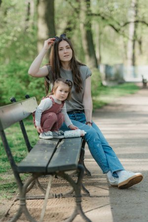 Téléchargez les photos : Une mère repose près de sa petite fille sur le banc dans le parc verdoyant. Une petite fille en velours joue avec sa mère dans le jardin. - en image libre de droit