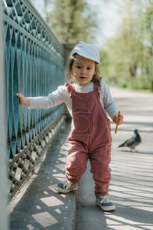 Photo for A female toddler eats a sausage close to the garden bridge fence. A baby girl in a cap and velvet overall walks near the pigeon in the park. - Royalty Free Image