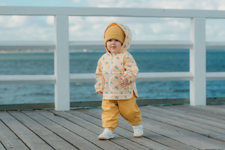 Photo for A beautiful toddler in a yellow jacket and pants strolls on the pier. A baby girl in the hood is having fun near the Baltic Sea. - Royalty Free Image