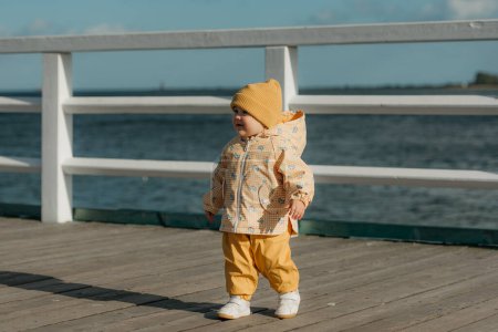 Foto de Un niño sonriente con una chaqueta amarilla y pantalones pasea por el muelle. Una niña se está divirtiendo cerca del Mar Báltico. - Imagen libre de derechos