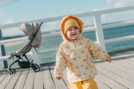 Foto de Un niño curioso en una chaqueta amarilla y pantalones pasea con un cochecito de bebé en el fondo en el muelle. Una niña en el capó se divierte cerca del mar Báltico. - Imagen libre de derechos