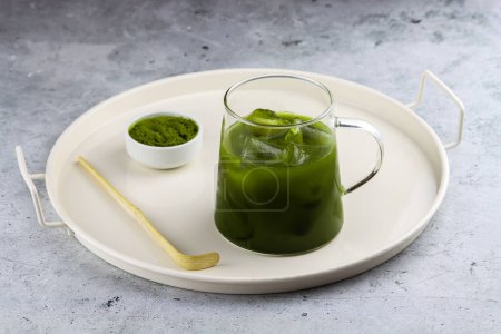 Antioxidant matcha green tea made of ground powder of green tea leaves in a transparent cup with ice cubes. Serving tray with glass of matcha, small bowl of matcha powder and matcha spoon on the table. Iced matcha tea on a serving tray