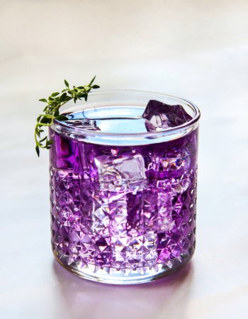 Photo for Purple drink made of violet liquor in a transparent drinking glass with ice cubes and a thyme sprig on the top. Violet cocktail. Iced colorful drink close up, isolated on a light grey blurred background in natural counterlight - Royalty Free Image