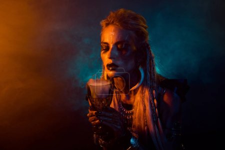 Foto de la mitología del norte mago vikingo niña mano mantenga poción taza naranja azul luces niebla aislado sobre fondo oscuro.