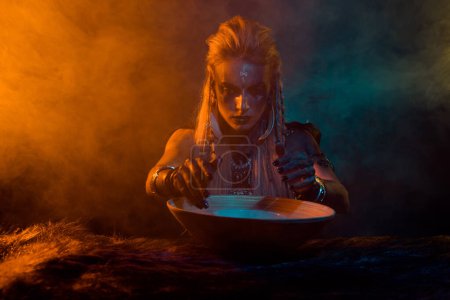 Photo of creepy viking comjurer woman demonic ritual potion bowl orange light foggy mist isolated on dark background.