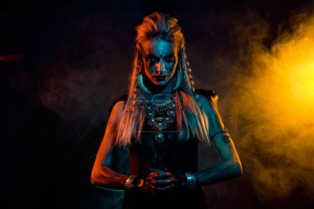 Portrait of fearless savage viking girl make up jewelry yellow blue lights foggy mist isolated on black background.