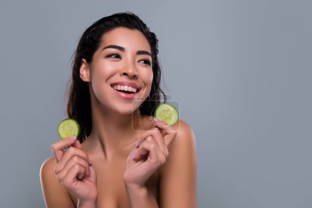 Téléchargez les photos : Photo de superbe asiatique femme tenir concombre légumes bio spa salon épaules off isolé fond de couleur grise. - en image libre de droit