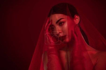 Téléchargez les photos : Photo de fille asiatique femme fatale toucher visage dentelle voile robe mariage épaules off isolé fond de couleur rouge foncé. - en image libre de droit