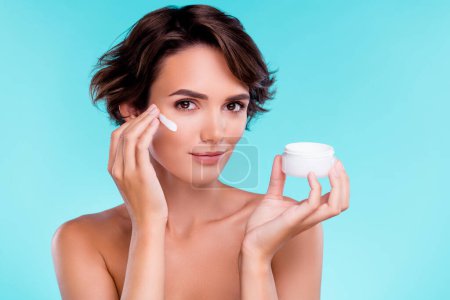 Foto de Foto de la hermosa dama suave aplicación de crema en las mejillas faciales para la piel sedosa suave sobre fondo azul. - Imagen libre de derechos