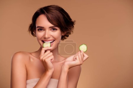Téléchargez les photos : Photo de joyeux joyeux jolie fille manger tranches de concombre profiter lifting du visage pour une procédure impeccable sur fond beige. - en image libre de droit
