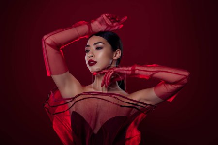 Téléchargez les photos : Photo de superbe asiatique femme fatale toucher visage posant modèle épaules off isolé rouge foncé couleur fond. - en image libre de droit