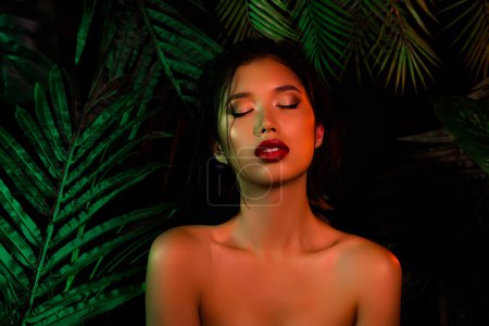Foto de Foto de chica japonesa vietnamita sobre hojas de palma verde oscuro cerrando los ojos con maquillaje decorativo. - Imagen libre de derechos