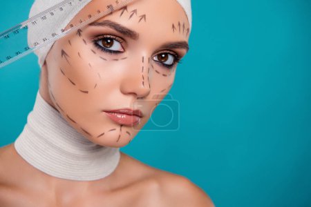 Foto de Foto de una paciente de cirugía plástica usando regla para examinar las proporciones de la cara sobre el fondo de color cian. - Imagen libre de derechos