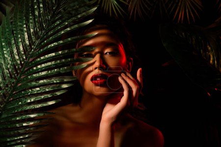 Téléchargez les photos : Photo de fille japonaise chinoise se cacher derrière les palmiers tropicaux toucher la peau sur fond sombre. - en image libre de droit