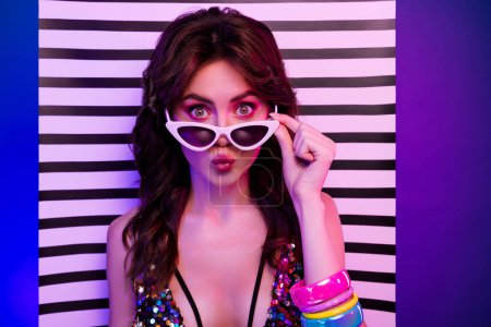 Photo portrait of pretty young girl amazed take off sunglass isolated on retro striped neon light background.