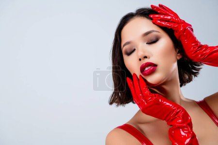Foto von atemberaubenden jungen Dame berühren Gesicht geschlossenen Augen tragen rotes Lederkleid isoliert auf hellgrauem Hintergrund.