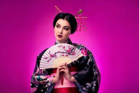 Téléchargez les photos : Portrait d'une magnifique geisha debout avec ventilateur dans ses mains sur la performance du théâtre sur fond de couleur rose. - en image libre de droit