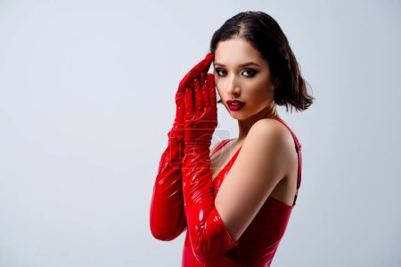 Foto de Foto de mujer joven atractiva mirada cara táctil cámara vestida ropa de cuero rojo aislado sobre fondo gris claro. - Imagen libre de derechos