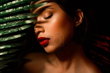 Téléchargez les photos : Gros plan photo de asiatique fille cacher visage sur vert exotique plante feuille avec les yeux fermés maquillage. - en image libre de droit