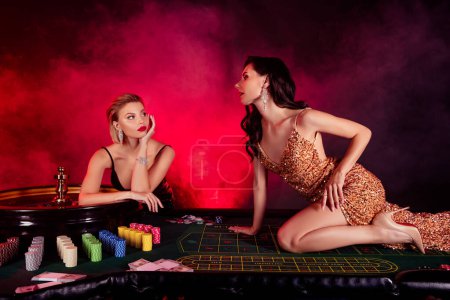 Téléchargez les photos : Photo de deux jolies dames excitées portent des robes sexy profiter de jouer à la roulette ensemble isolé fond de brouillard rouge. - en image libre de droit