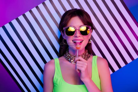 Photo for Photo of happy girl lick tasty lolipop stand over striped wall isolated neon multicolor background. - Royalty Free Image