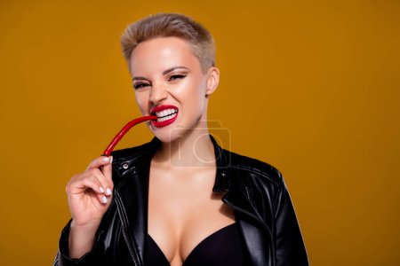 Foto de Retrato de la joven enojada comiendo pimiento rojo picante vestido sostén y chaqueta sobre fondo de color marrón pastel. - Imagen libre de derechos