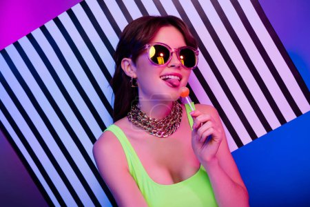 Foto de Photo of happy girl in sunglass gold jewelry top lick lolipop over striped banner isolated pink neon background. - Imagen libre de derechos