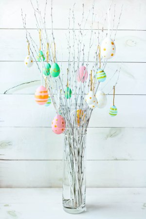 Foto de The branches of the trees decorated with multi-colored Easter eggs stand in a vase on a white wooden background. Easter tree. Easter composition. - Imagen libre de derechos