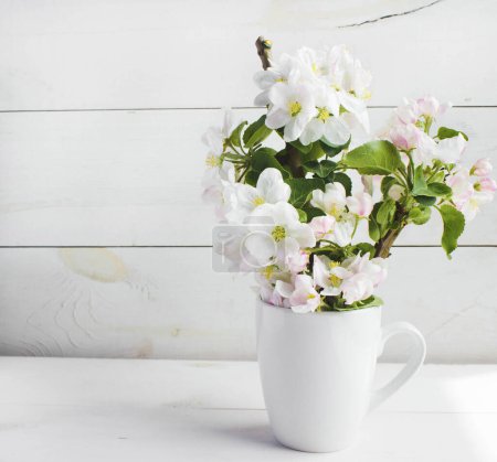 A beautiful sprig of an apple tree with white flowers in a cap against a white wooden background. Blossoming branch in a glass with water. Spring still life. Place for text. Concept of spring or mom day.