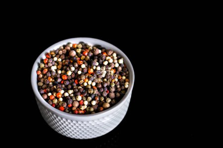 A mixture of pepper varieties with peas on the white plate on the black background. Heap of various pepper. Mix of red, black and white peppercorn seeds isolated on white. Top view.
