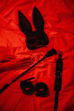 Foto de Set of BDSM accessories with female hands is lying on bed. Flogger whip, handcuffs, mask and choker for sex with submission and domination - Imagen libre de derechos