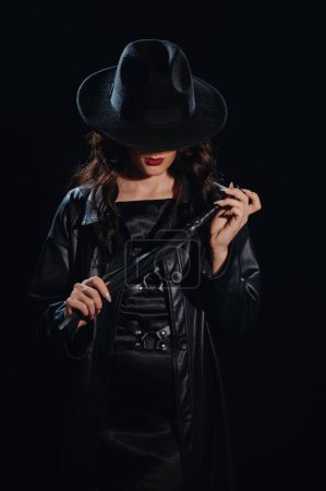 Téléchargez les photos : Sexy maîtresse dominante féminine avec un fouet pour le sexe BDSM avec soumission et domination dans un imperméable en cuir et un chapeau dans le style noir - en image libre de droit
