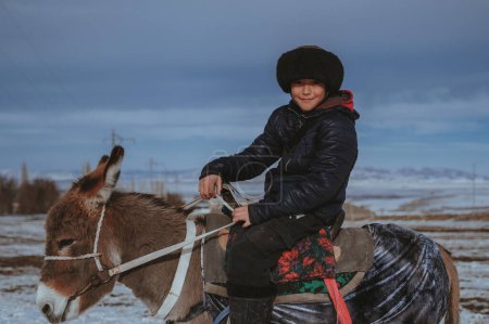 Foto de TAU SAMALY, KAZAKHSTAN - 23 de diciembre de 2023: Niño kazajo montado en un burro en invierno en un campo cerca de Tau Samaly en Kazajstán - Imagen libre de derechos