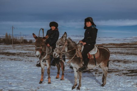 Foto de TAU SAMALY, KAZAKHSTAN - 23 de diciembre de 2023: dos amigos kazajos niños montados en un burro en invierno en un campo cerca de Tau Samaly en Kazajstán - Imagen libre de derechos