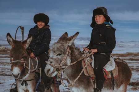 Foto de TAU SAMALY, KAZAKHSTAN - 23 de diciembre de 2023: dos amigos kazajos niños montados en un burro en invierno en un campo cerca de Tau Samaly en Kazajstán - Imagen libre de derechos
