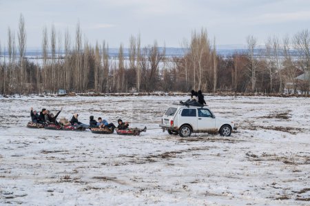 Téléchargez les photos : TAU SAMALY, KAZAKHSTAN - 23 DÉCEMBRE 2023 : des enfants kazakhs heureux montent des tubes extrêmes derrière une voiture dans un champ enneigé en hiver au centre récréatif Tau Samaly au Kazakhstan - en image libre de droit