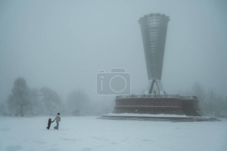 Téléchargez les photos : SHYMKENT, KAZAKHSTAN - 19 DÉCEMBRE 2023 : Une mère et un fils traversent le Parc de l'Indépendance à Shymkent au Kazakhstan au Monument Altyn Shanyrak en hiver avec de la neige et du brouillard - en image libre de droit