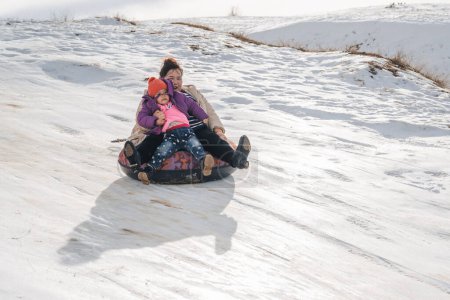 Téléchargez les photos : TAU SAMALY, KAZAKHSTAN - 23 DÉCEMBRE 2023 : une famille kazakhe heureuse descend un toboggan en hiver avec de la neige au centre récréatif Tau Samaly au Kazakhstan - en image libre de droit