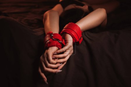 Foto de Manos femeninas en cuero BDSM esposas para sumisión y dominación sexo en una sábana negra en primer plano. Mujer sumisa esclava chica acostada en la cama en el dormitorio - Imagen libre de derechos