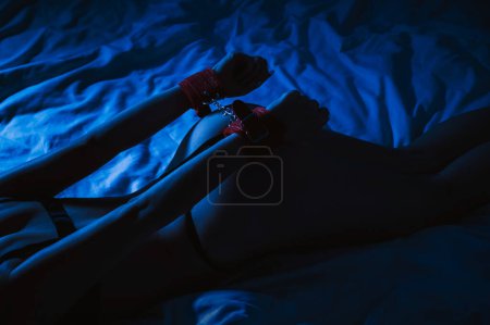 Téléchargez les photos : Submissive woman girl with a sexy body and ass in panties with handcuffed hands lying on bed in bedroom at night. BDSM sex with submission and domination with blue neon light - en image libre de droit