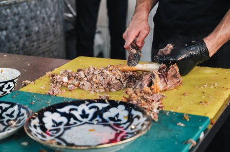 male chef cuts boiled meat from a lamb bone with a knife to cook traditional Uzbek pilaf in the kitchen at a restaurant in Uzbekistan