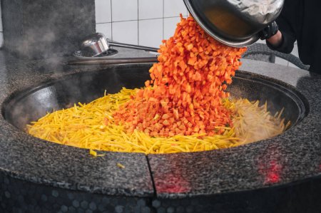 un chef masculin ajoute des carottes à un chaudron pour cuisiner du pilaf ouzbek traditionnel dans la cuisine d'un restaurant en Ouzbékistan