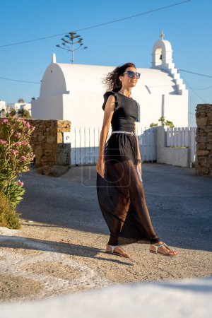 Foto de Mujer joven en vestido negro largo al lado de una iglesia griega - Imagen libre de derechos