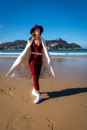 Stilvolle Frau mit kastanienbraunem Kleid und weißem Mantel an der Playa de la Concha, San Sebastian