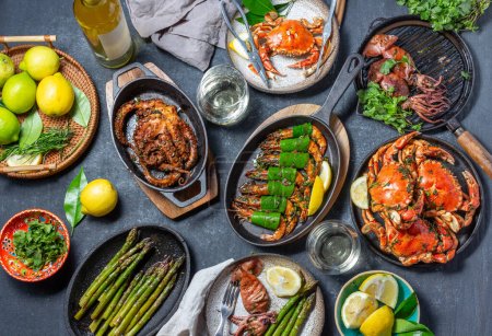 Photo for Set table with seafood dishes - cooked crabs, tiger shrimps, grilled octopus and squids on cast iron grilled pans and plates, White wine. Top view. - Royalty Free Image