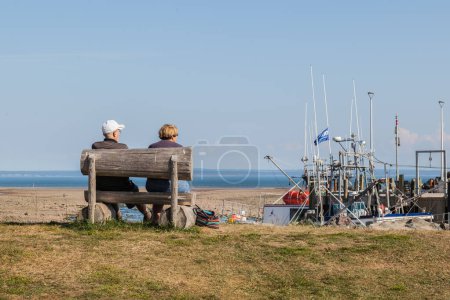 Téléchargez les photos : VILLAGE D'ALMA, NB, CANADA - LE 4 AOÛT 2017 : Vieux couple assis sur le banc de bois face à la rivière dans le village de pêcheurs d'Alma sur la baie de la Fundy, NB - en image libre de droit