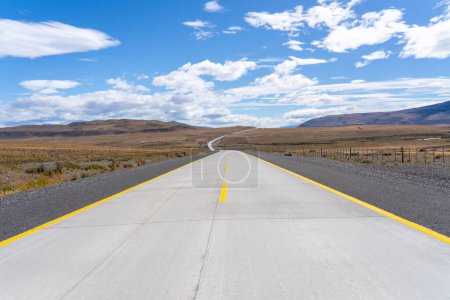A long straight road with yellow lines leading towards mountains that crosses the Atacama desert in Chile. Sun with clouds in the blue sky.