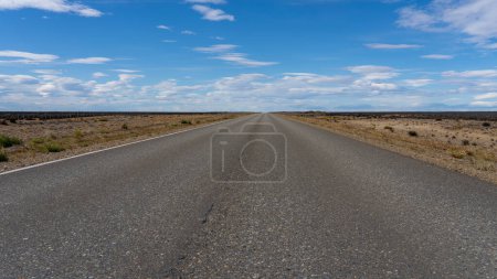 Eine lange, gerade Straße ohne Linien, die durch die Atacama-Wüste in Chile führt. Sonne mit Wolken am blauen Himmel.