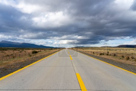 A long straight road with yellow lines leading towards mountains that crosses the Atacama desert in Chile on a dark cloudy day. 